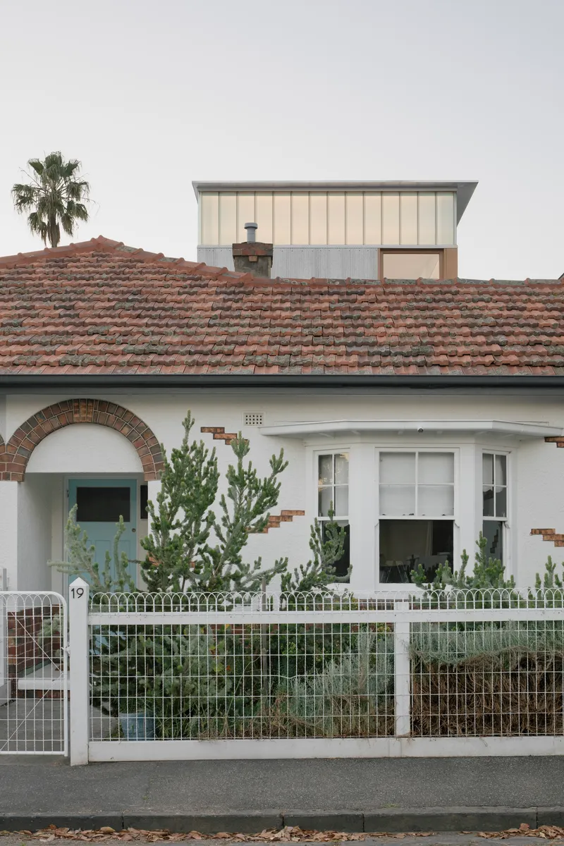 Street view of the white rendered bungalow with terracotta tile roof and metal-clad addition rising behind
