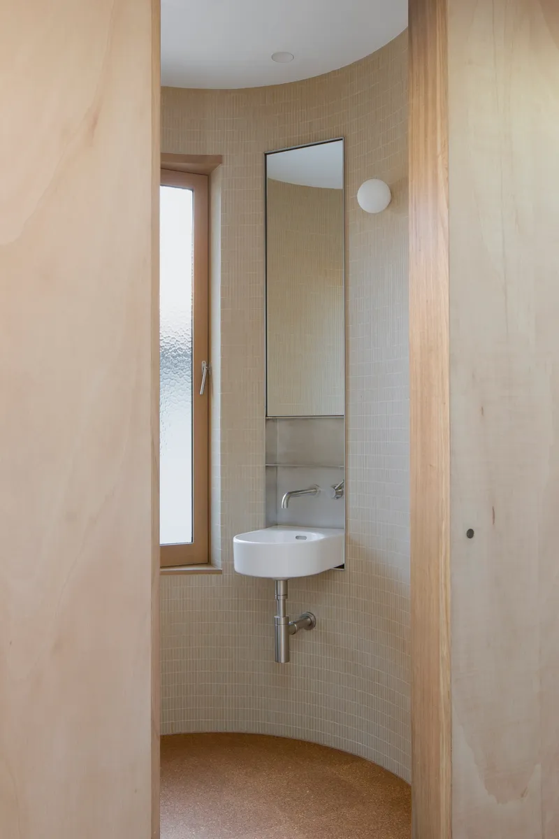 Curved powder room with pale mosaic tile walls and a wall-mounted basin beneath a vertical mirror