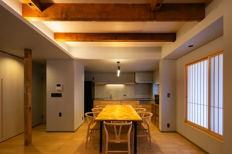 Dining area with exposed timber beams, slatted window screen and kitchen beyond in warm evening light