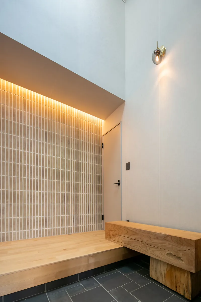 Entry vestibule with grey rectangular tile wall, recessed uplighting, floating timber bench and dark slate floor