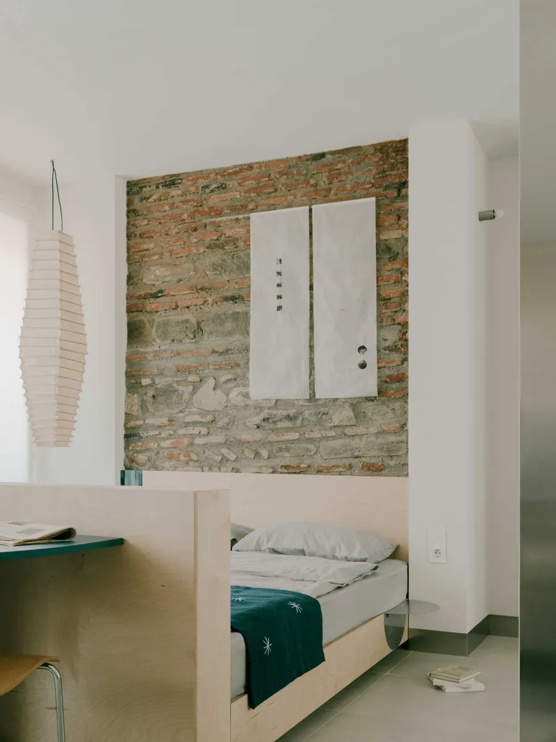 View across plywood platform bed to exposed brick feature wall with mounted white door panel