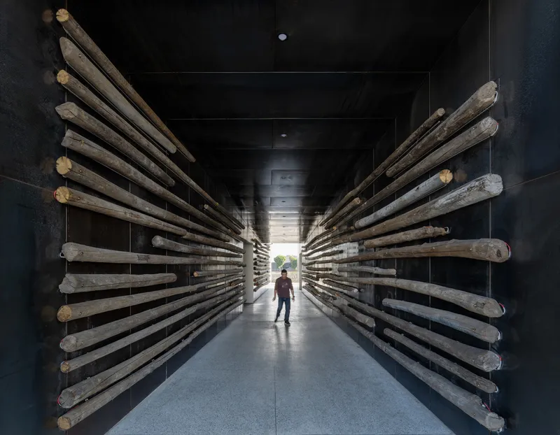 Corridor lined with reclaimed timber poles projecting from black walls as a visitor walks toward daylight