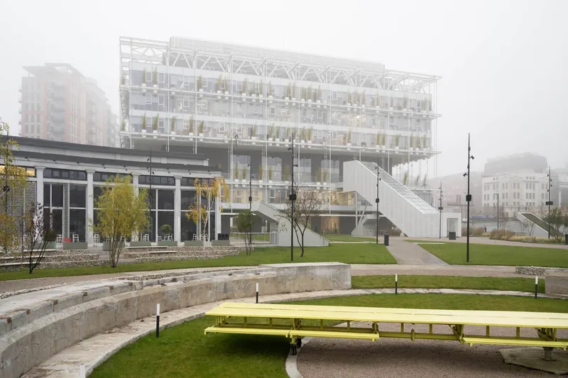 Terraced public park with yellow benches and concrete amphitheater steps leading toward the glass building