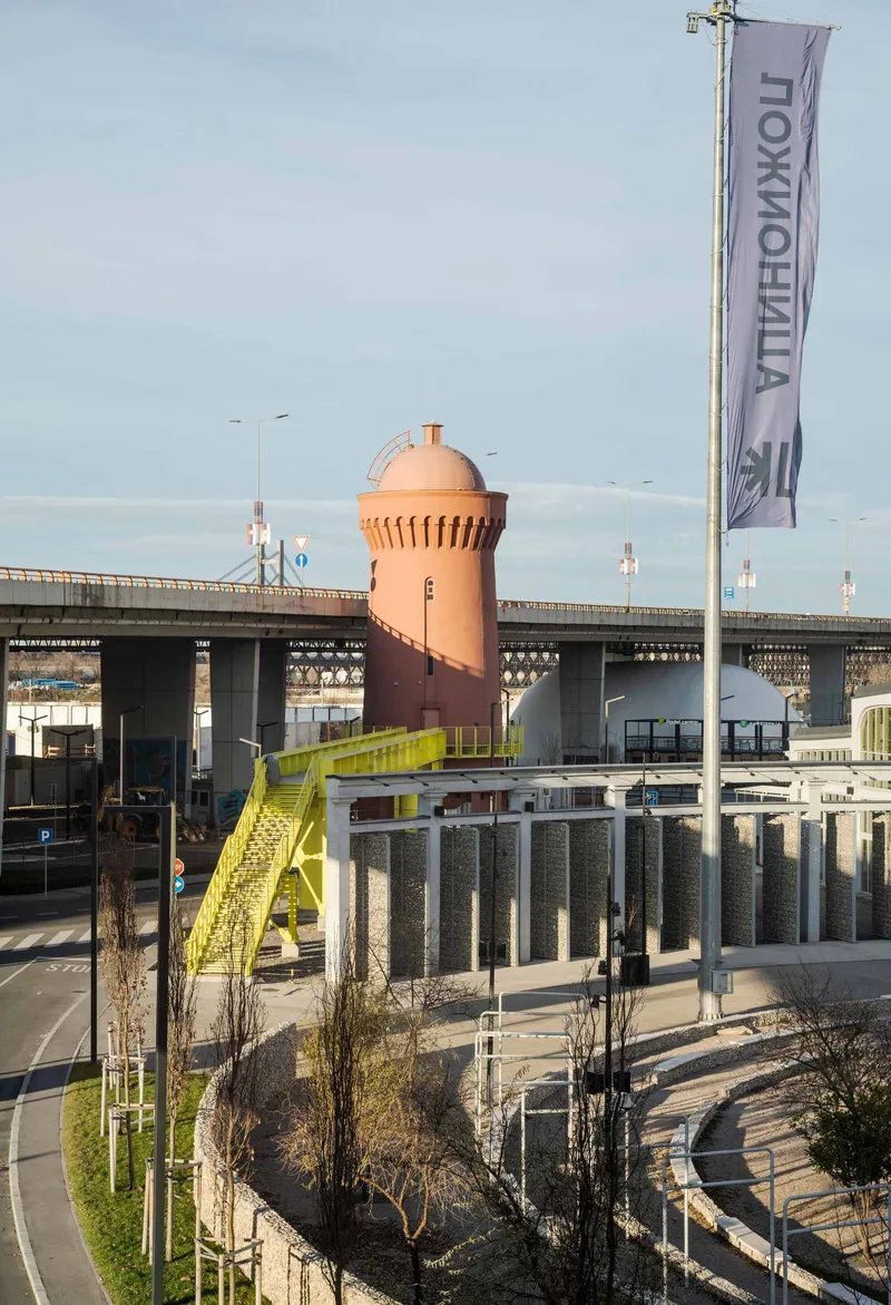 Cylindrical tower with corrugated terracotta cladding and domed roof beside a highway overpass at dusk