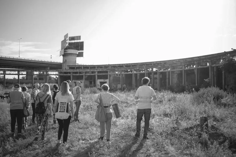 Group of visitors walking through overgrown grasses towards a curved colonnade and control tower at dusk