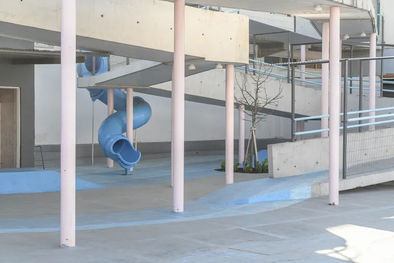 View through pink columns showing blue spiral slide and young planted tree with wire mesh railings