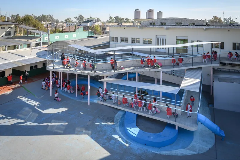 Elevated courtyard with blue tubular slide and schoolchildren in red uniforms gathering beneath corrugated metal canopies