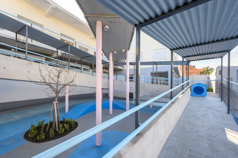 Concrete ramp with pink cylindrical columns and corrugated metal canopy shading a planted circular bed