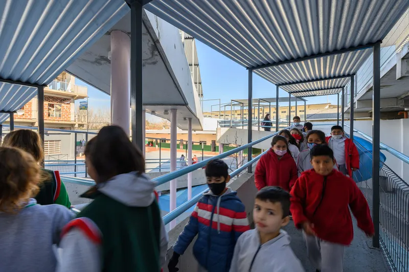 Students walking along a covered ramp with wire mesh railings under corrugated metal overhead canopy in afternoon light