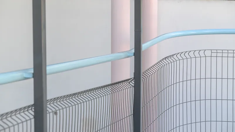 Close-up of curved wire mesh fence meeting blue metal handrail with pink columns in background