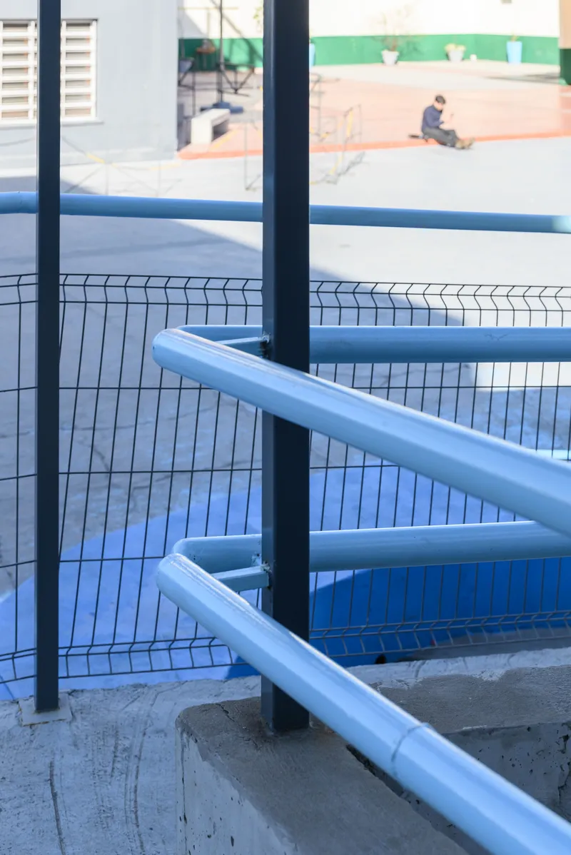 Blue tubular handrails and wire mesh fencing along a concrete staircase with a schoolyard beyond