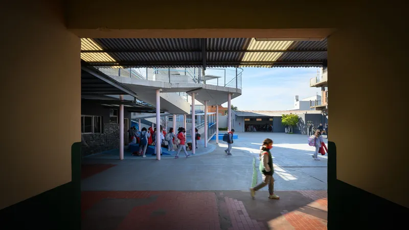 View through a shadowed corridor toward a sunlit courtyard with children walking and corrugated metal canopy