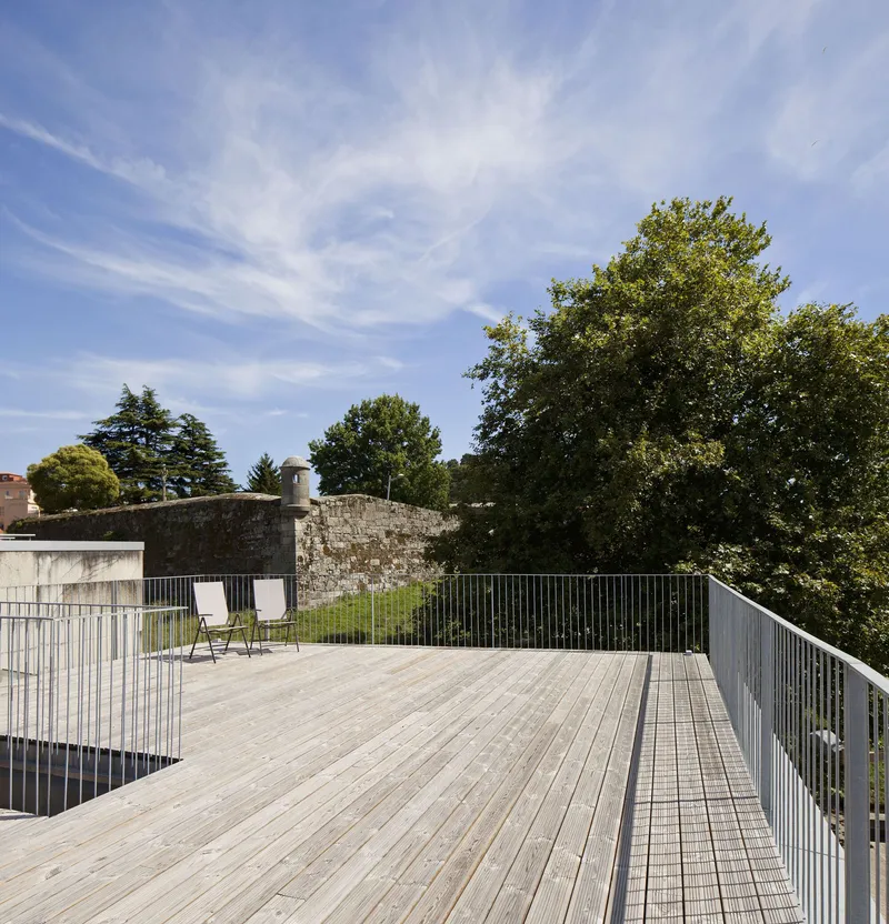 Timber deck terrace with metal mesh railings beside a stone wall and mature trees under open sky