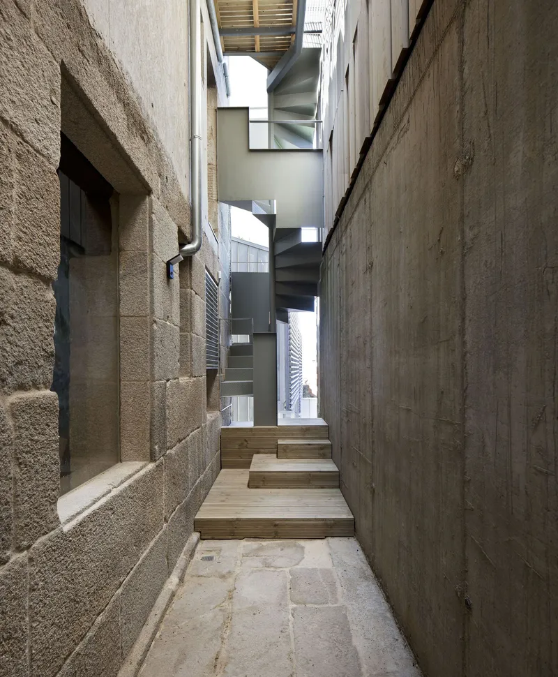 Narrow passage between exposed concrete walls with timber stairs ascending toward daylight above