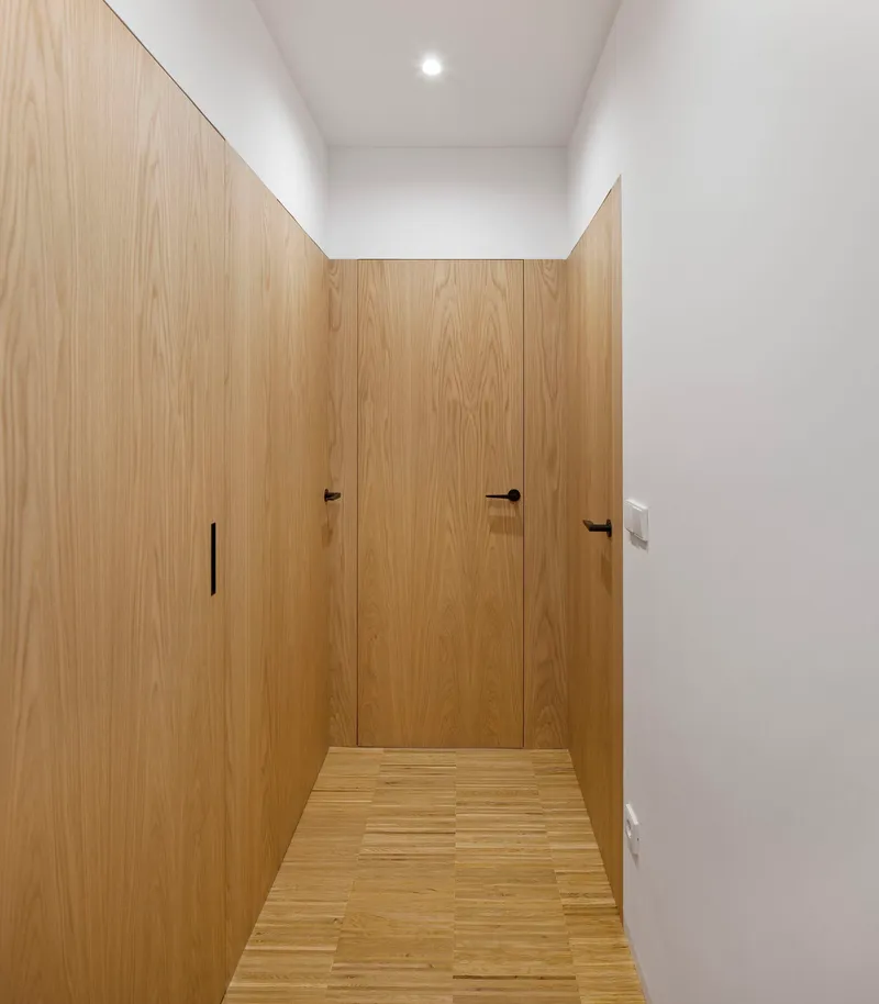 Corridor lined with flush oak veneer panels concealing doors and storage under recessed ceiling light