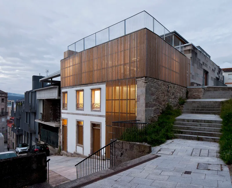 Corner facade showing timber and corrugated metal volumes rising above a stone retaining wall at twilight