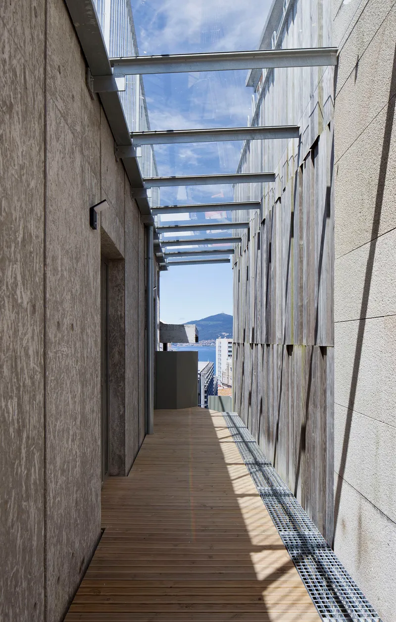 Timber deck corridor with overhead glass canopy between wood-clad and concrete walls framing a harbor view