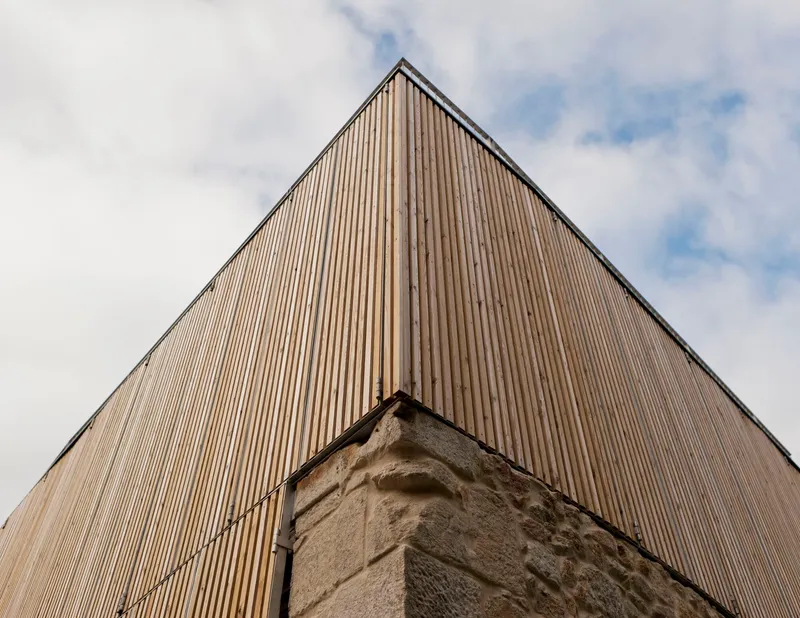 Upward view of the vertical timber cladding meeting an existing stone corner under cloudy skies