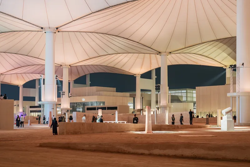 Evening view of the covered plaza with illuminated stone service counters and visitors beneath tensile canopies