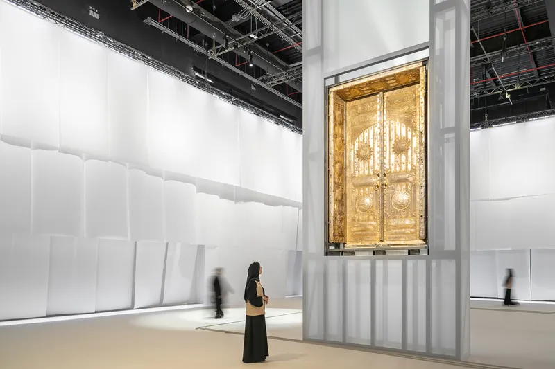 Gallery space with white quilted walls displaying ornate gilded doors on a raised platform with visitors