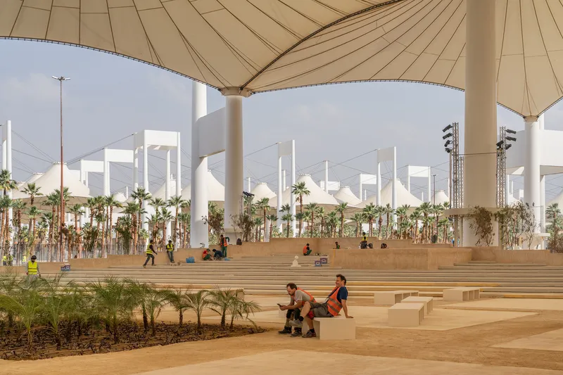 Tiered stone seating with planted palm trees and people resting beneath the white column and canopy structure