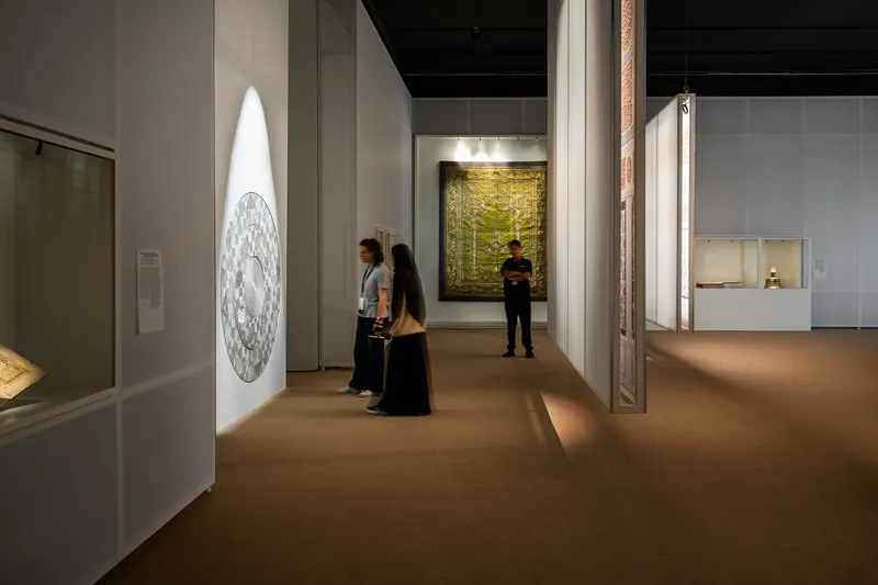 Gallery corridor with visitors viewing illuminated artworks on white walls and glass display cases