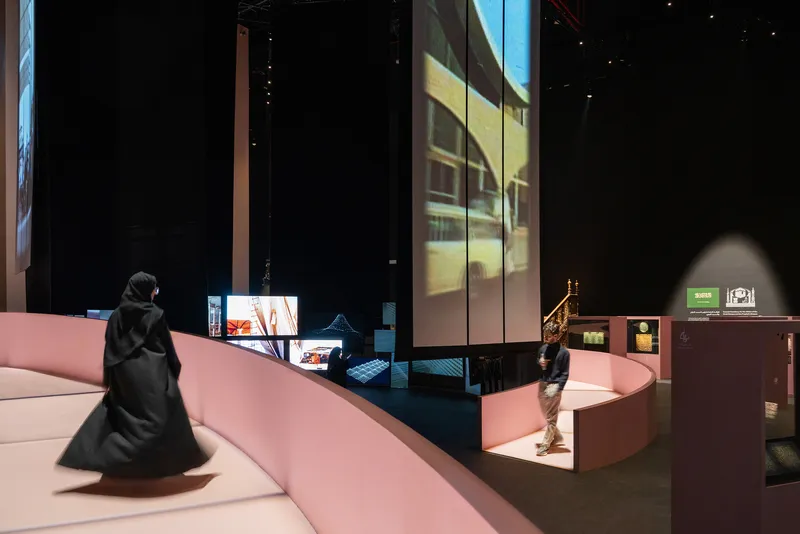 Curved pink platform with visitors and interactive displays beneath projected video on gallery walls
