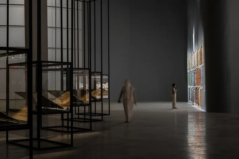 Gallery hall with black steel frame shelving and visitors near illuminated textile hanging on wall