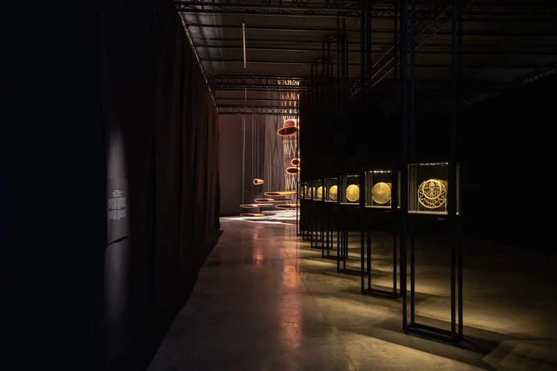 Exhibition corridor with backlit artifact cases and suspended copper lighting elements under exposed ceiling infrastructure