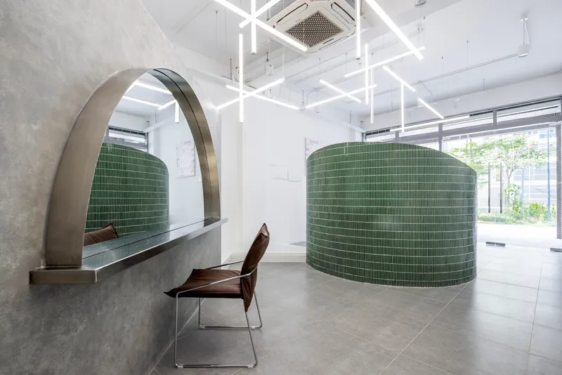Curved green tile wall with arched concrete canopy and a wooden chair near daylit storefront windows
