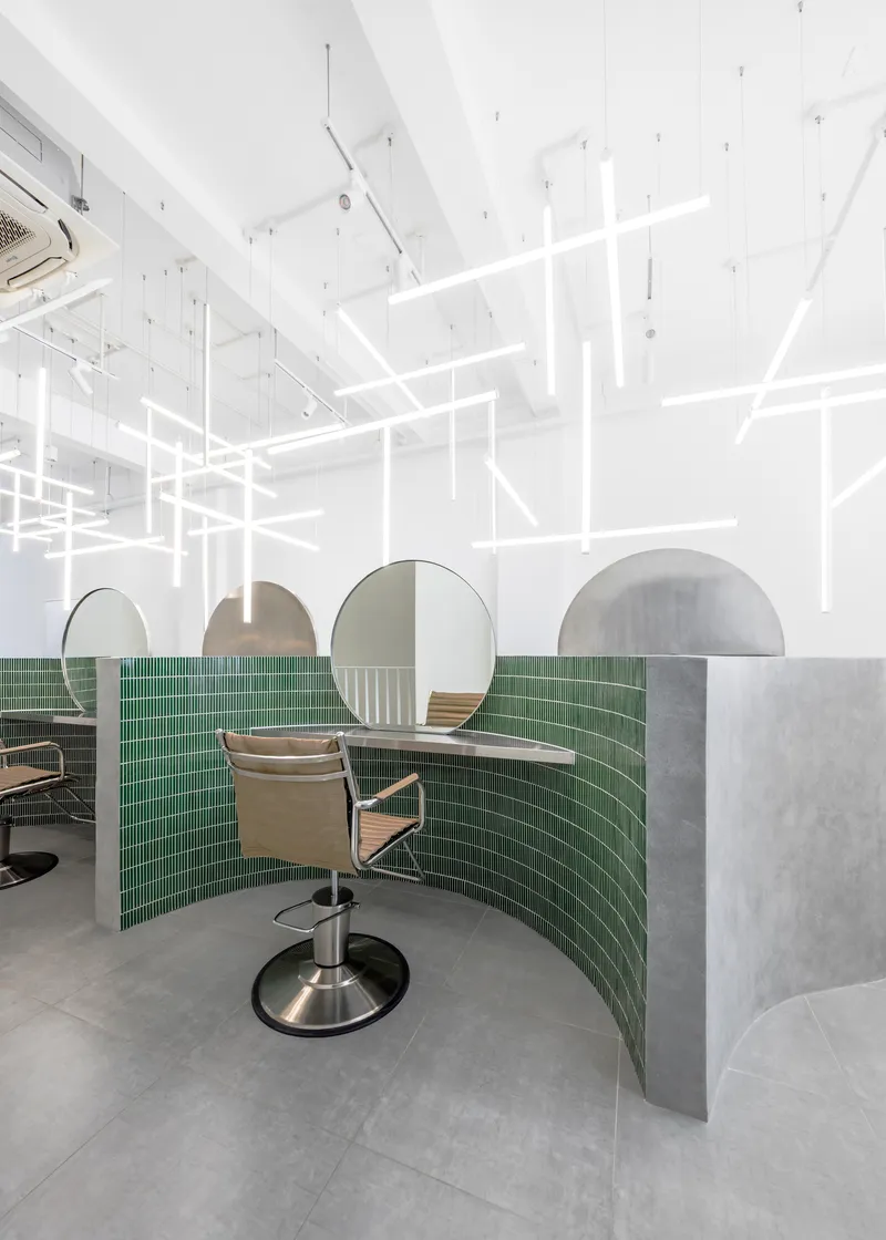 Salon station with curved green tile partition, circular mirror, and hydraulic chair under gridded ceiling lights