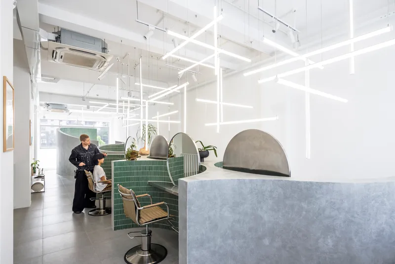 White grid light fixture suspended over green tiled salon stations with chrome chairs and a stylist at work