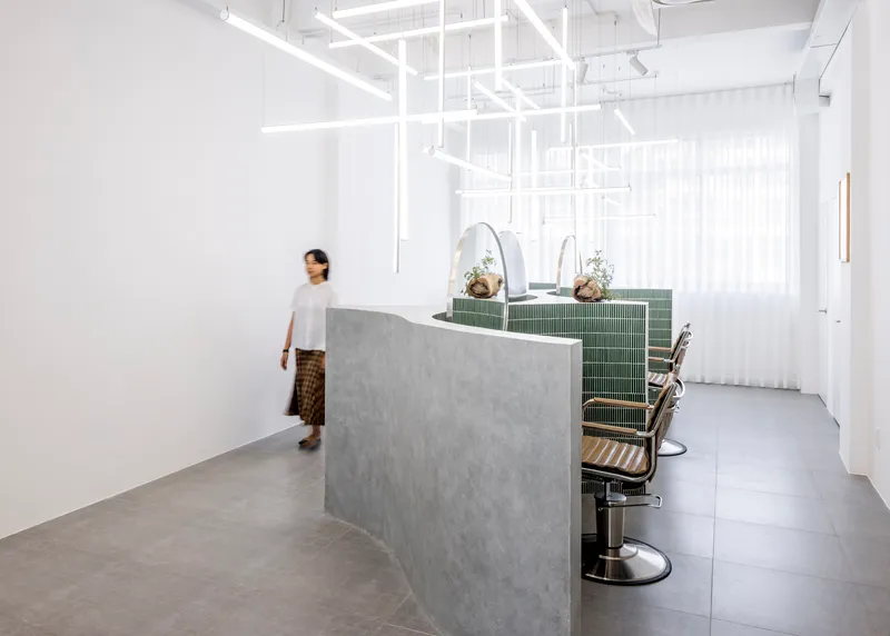 Person walking past curved concrete reception counter in white salon interior with suspended linear lights