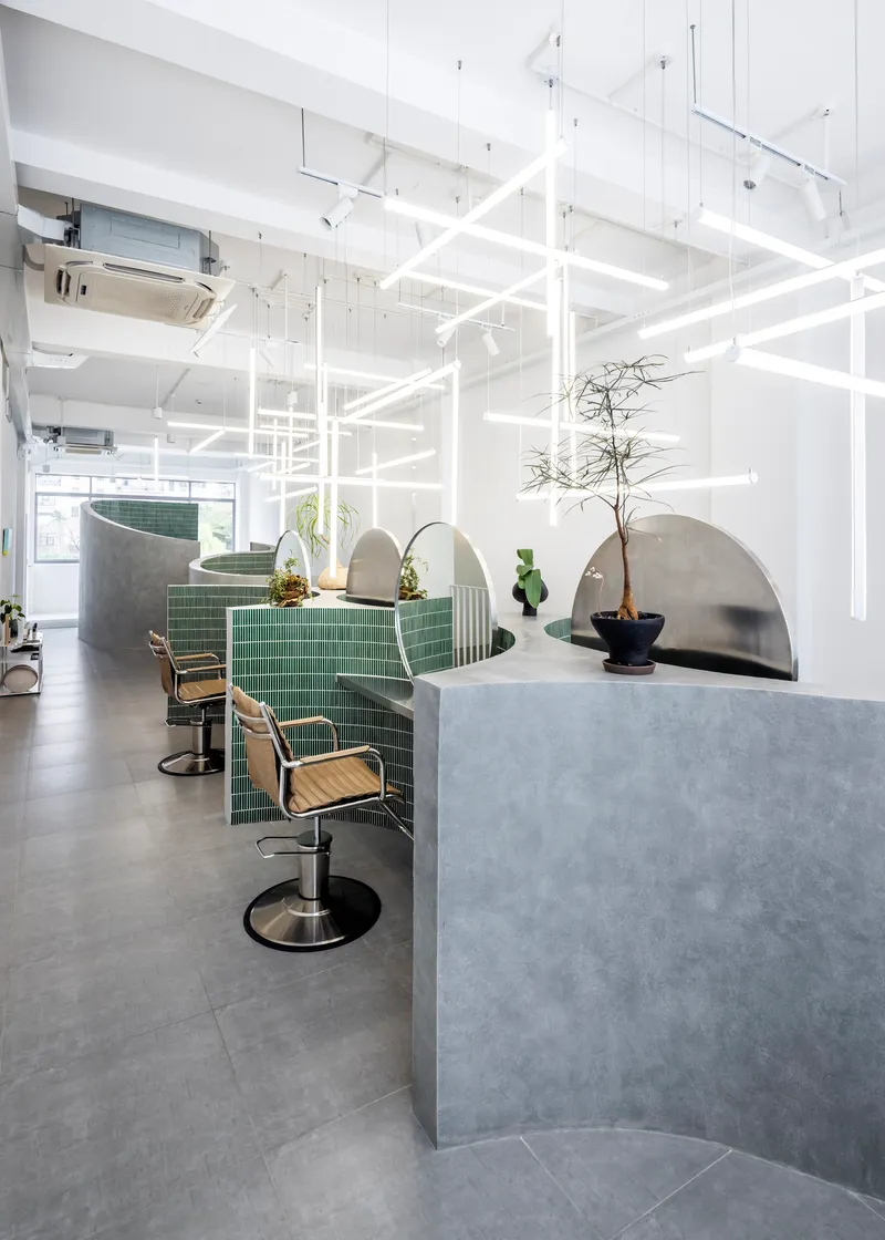 Salon workstations with green tile partitions, curved concrete screens and potted plants under grid lighting