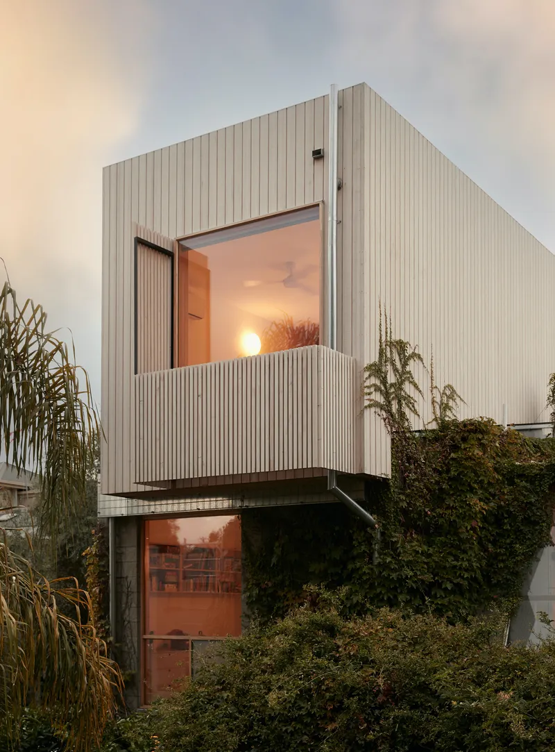 White vertical board cladding with cantilevered balcony opening at dusk with surrounding garden vegetation