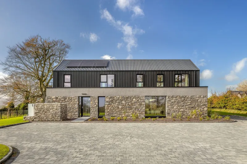 Front facade combining stone walls and dark corrugated cladding with rectangular windows under afternoon clouds