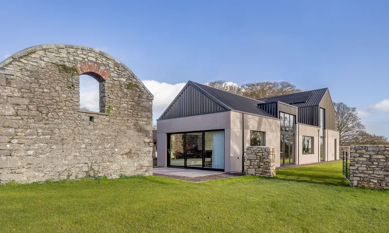 Stone barn ruin arch next to rendered and corrugated volumes on grass lawn