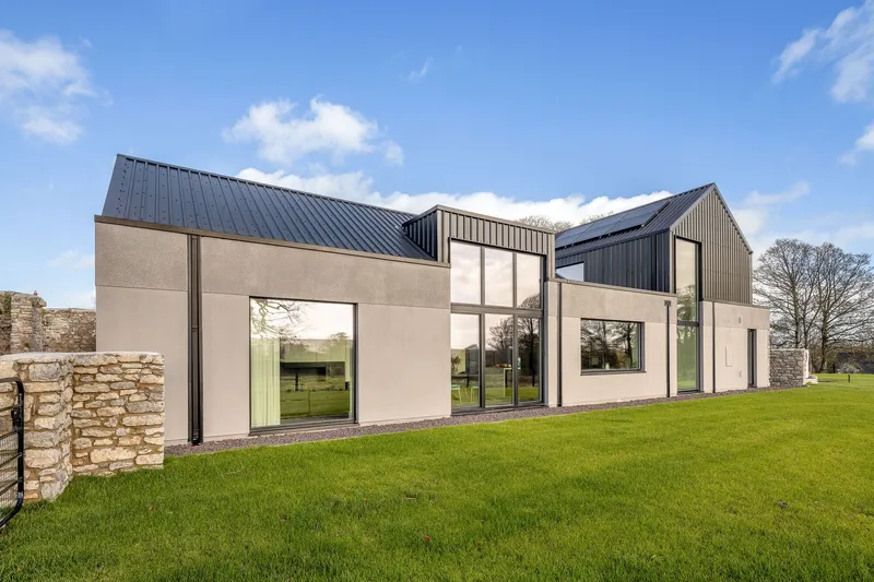 Rear facade with smooth render walls and corrugated metal roofs flanked by stone ruins and a lawn