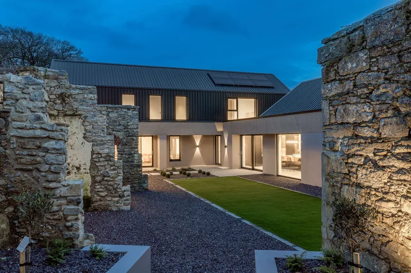 Gravel courtyard framed by illuminated stone ruins and the glazed facade at dusk