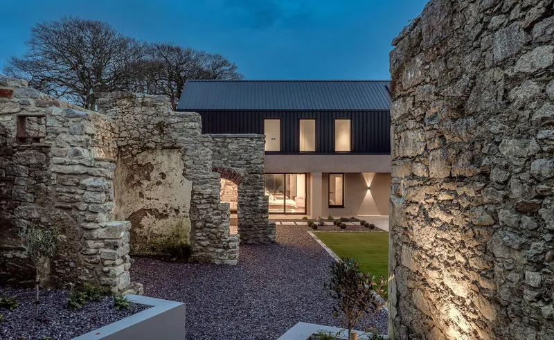 View through the ruined stone archway toward the lit windows and gravel courtyard at twilight