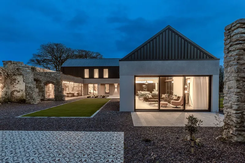Glass living pavilion opening to the courtyard with metal roofed volumes and stone walls at dusk
