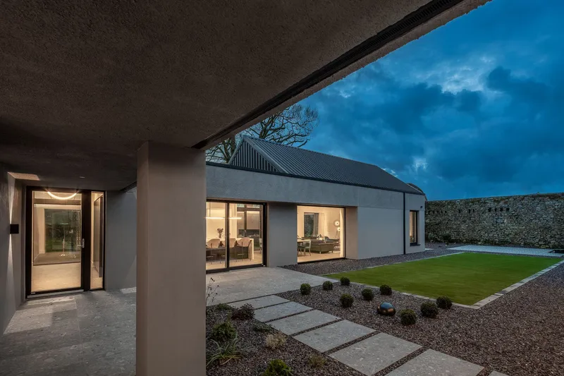 Covered terrace looking across paved stepping stones and gravel toward the illuminated interior at twilight