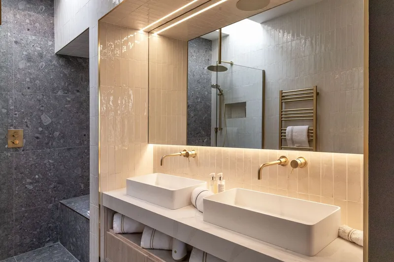 Bathroom vanity with dual vessel sinks, backlit vertical tiles, and brass fixtures beneath a timber-lined ceiling