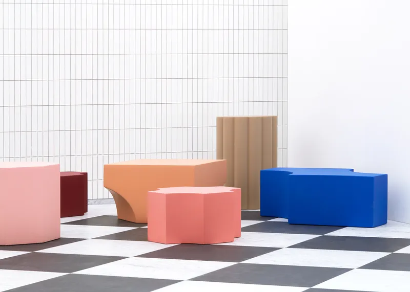 Colored seating blocks in pink, coral, and blue arranged on a chequered floor in bright sunlight