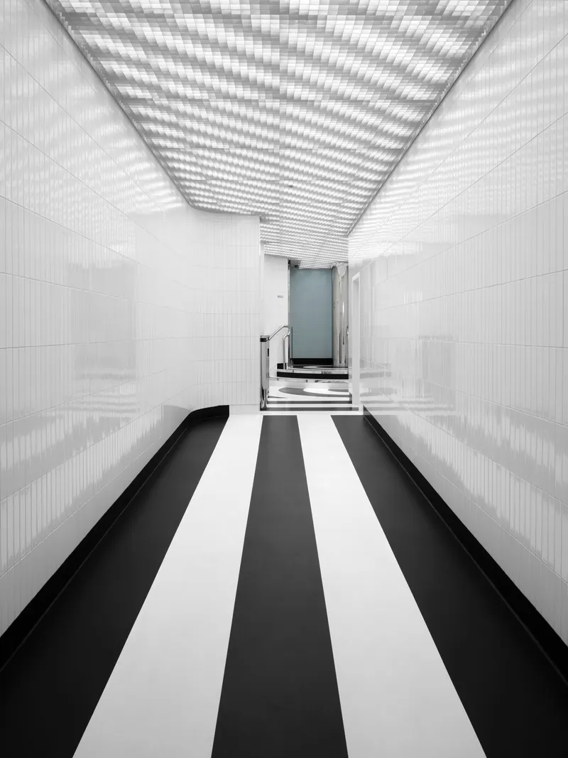 Corridor with black and white striped floor beneath a perforated ceiling casting shadow patterns