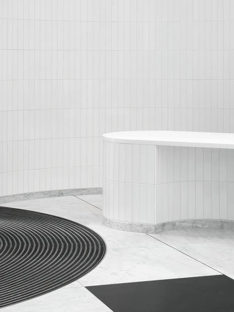 White cylindrical reception desk beside curved black floor mat on terrazzo in a tiled lobby