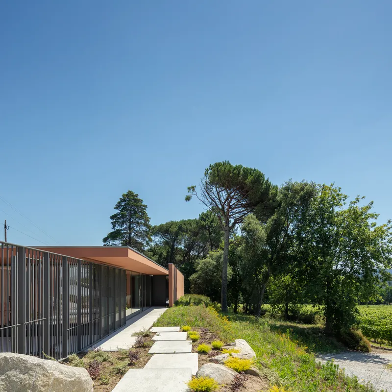 Concrete pathway bordered by planted beds leading to a terracotta-roofed building with vertical metal screening
