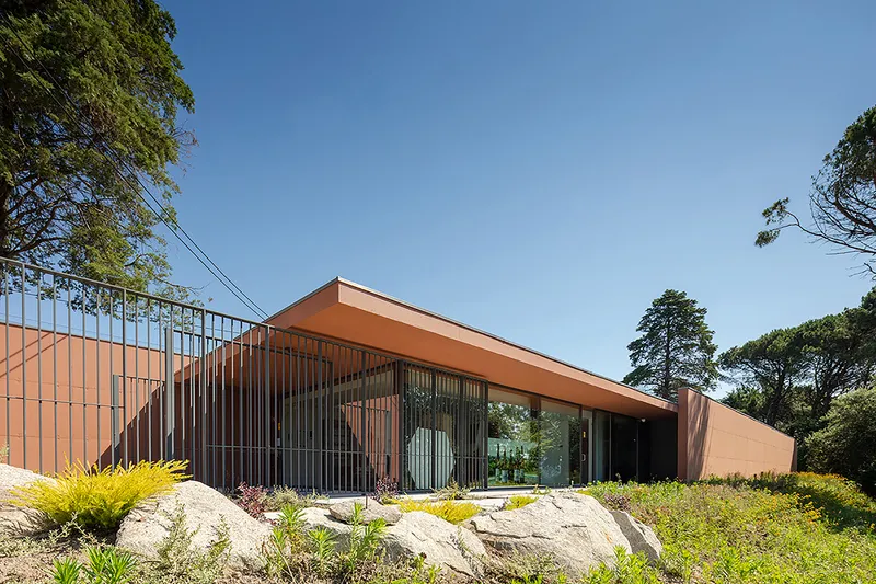 Angled view of the terracotta-clad volume with vertical screening and a landscaped boulder garden under blue sky