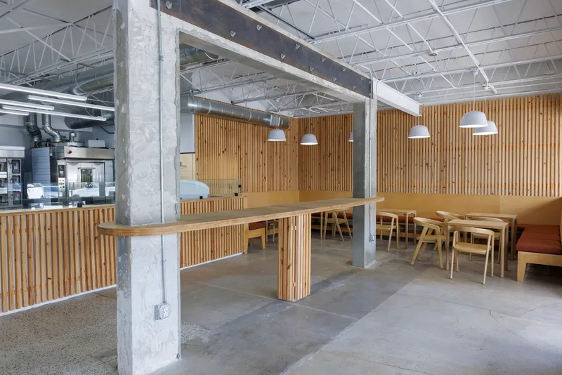 Long communal timber table wrapping around a concrete column with exposed truss ceiling above