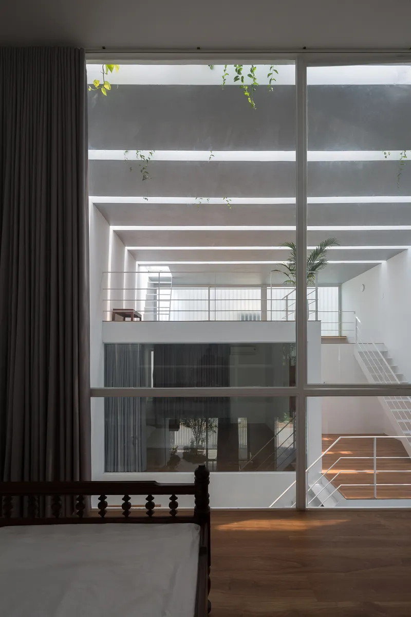 View from bedroom through floor-to-ceiling window into open interior atrium with potted palm and timber floors